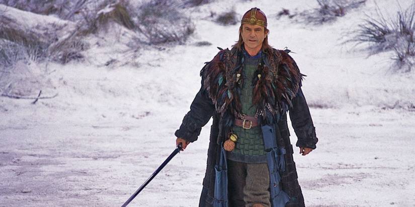 Sam Neill as Merlin wearing a feathered coat and holding a sword in the snow in 'Merlin'