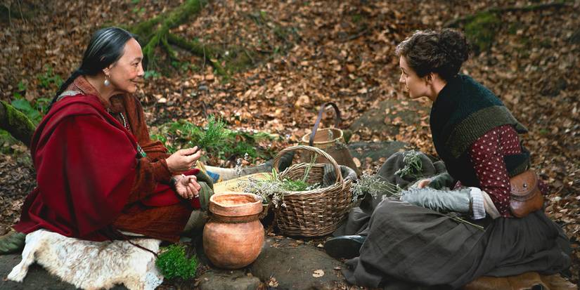 Adawehi sitting with Claire in the forest as the two women talk and gather herbs in Outlander Season 4