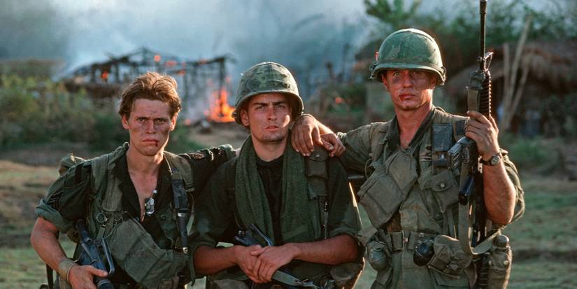 Charlie Sheen, Willem Dafoe, and Tom Berenger as soldiers look at the camera with a burning building behind in Platoon.