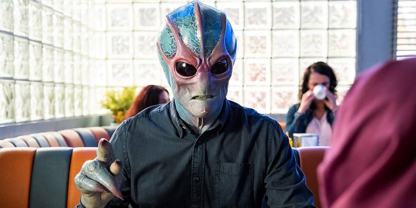 Alan Tudyk in alien form sitting at a diner in Resident Alien