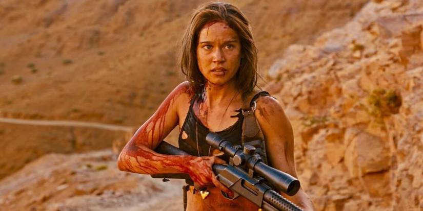 Matilda Lutz as Jen, covered in blood and standing out in the desert while holding a large gun in Revenge (2017)