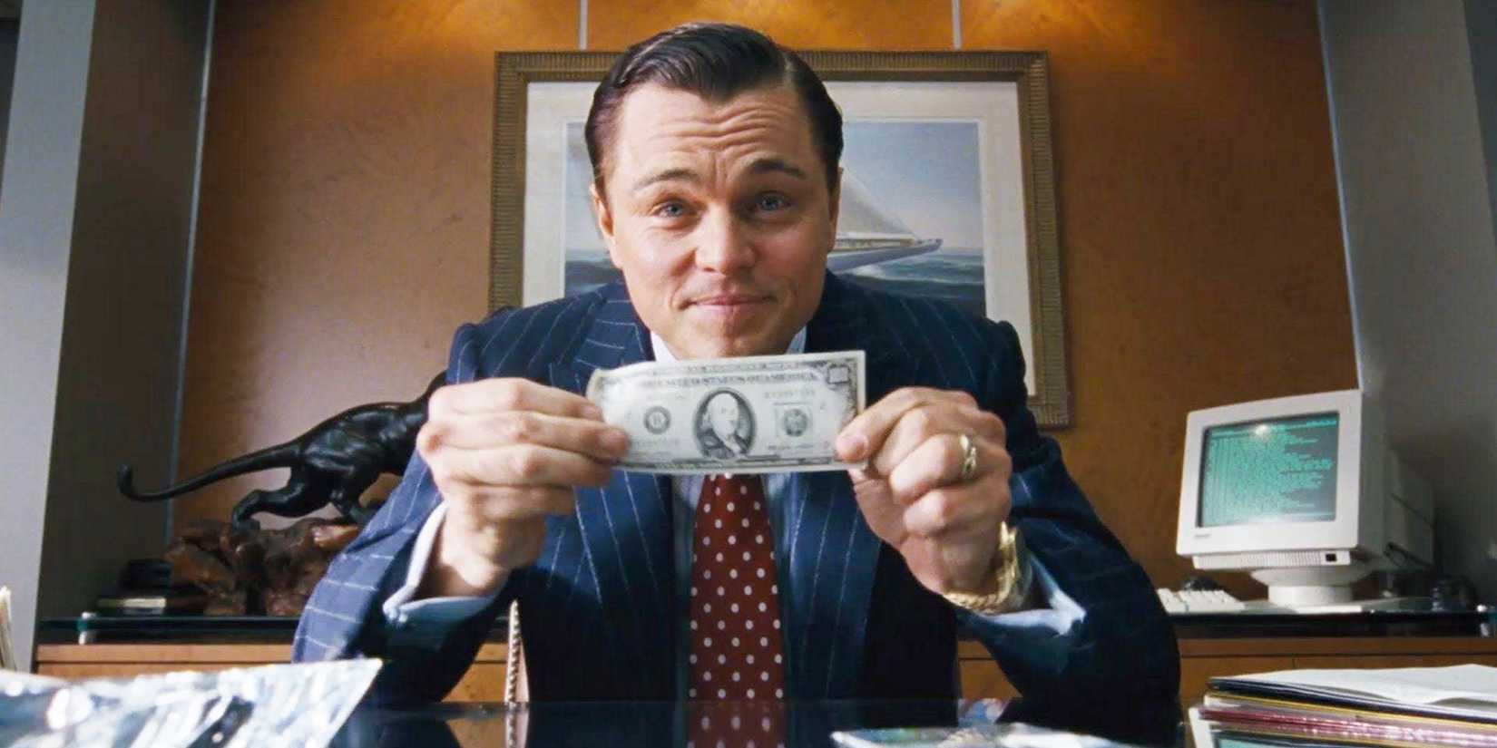 Leonardo DiCaprio as Jordan Belfort holding a bill while looking at a camera in The Wolf of Wall Street.