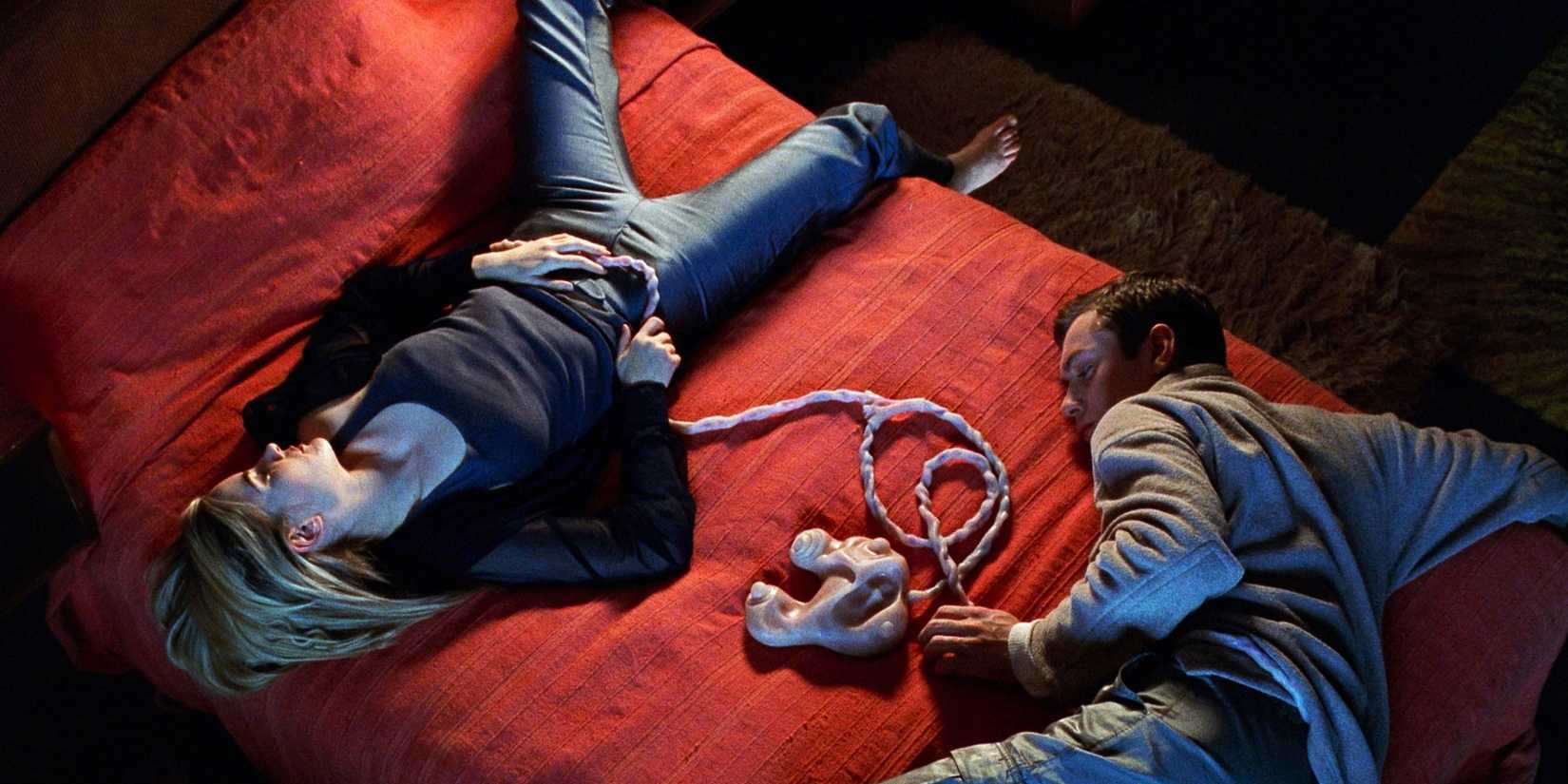 Jennifer Jason Leigh as Allegra and Jude Law as Ted lay on a bed with their umbi-cords connected to the game pod in eXistenZ 