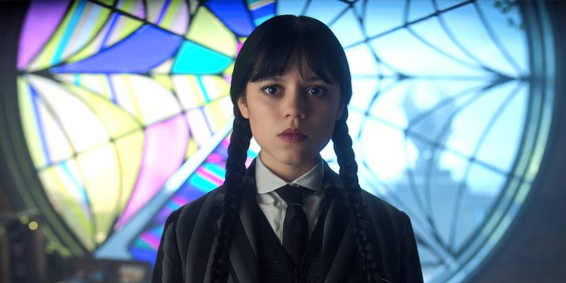 Jenna Ortega as Wednesday Addams in front of her and Enid's half-colored window in 'Wednesday'.