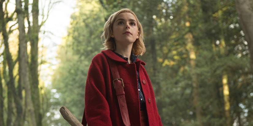 Kiernan Shipka as Sabrina Spellman in a red coat in the woods in Chilling Adventures of Sabrina.