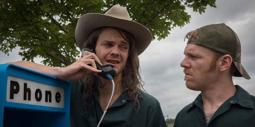 Jack Quaid talking on a payphone while Brian Gleeson looks at him, confused, in Logan Lucky