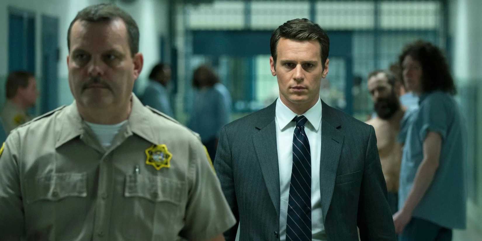 An agent in a suit and tie is escorted by a guard through a prison as inmates look on in Mindhunter.