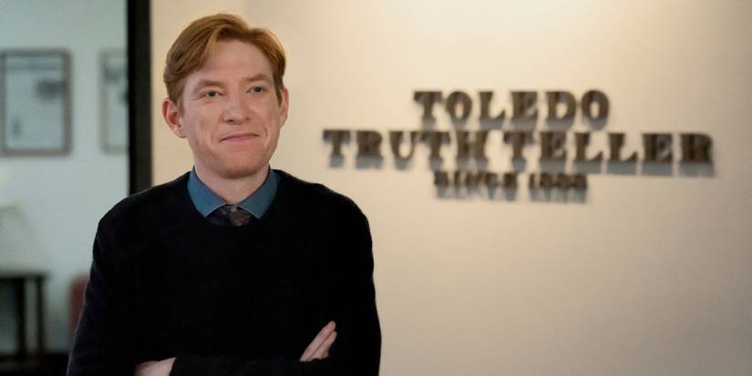 Domhnall Gleeson stāv sienas zīmju priekšā, kas lasāms "Toledo patiesības stāstītājs" papīrā. 