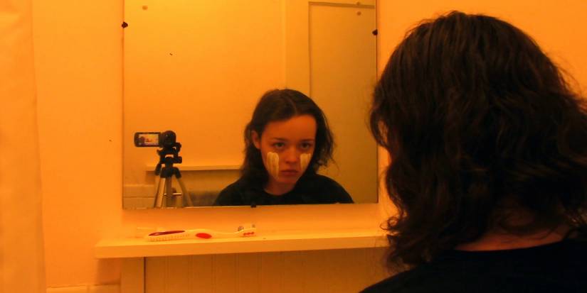 A young woman staring at her reflection in a mirror with a video camera beside her in We're All Going to the World's Fair (2021)
