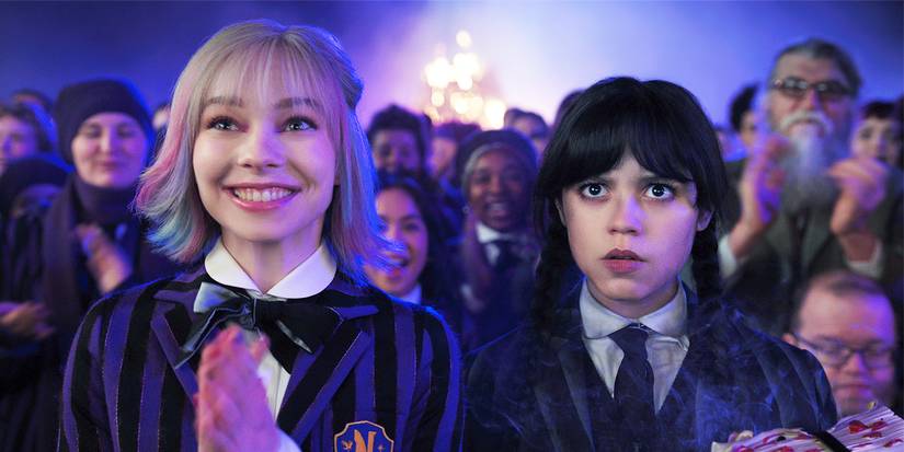 Emma Myers and Jenna Ortega staring ahead at a school event with a fire behind in Wednesday Season 2.