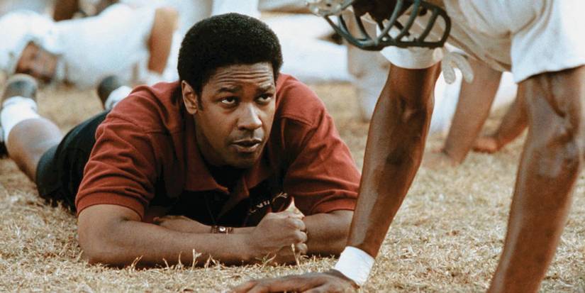 Denzel Washington lies on his stomach on the ground coaching a football player in Remember the Titans.