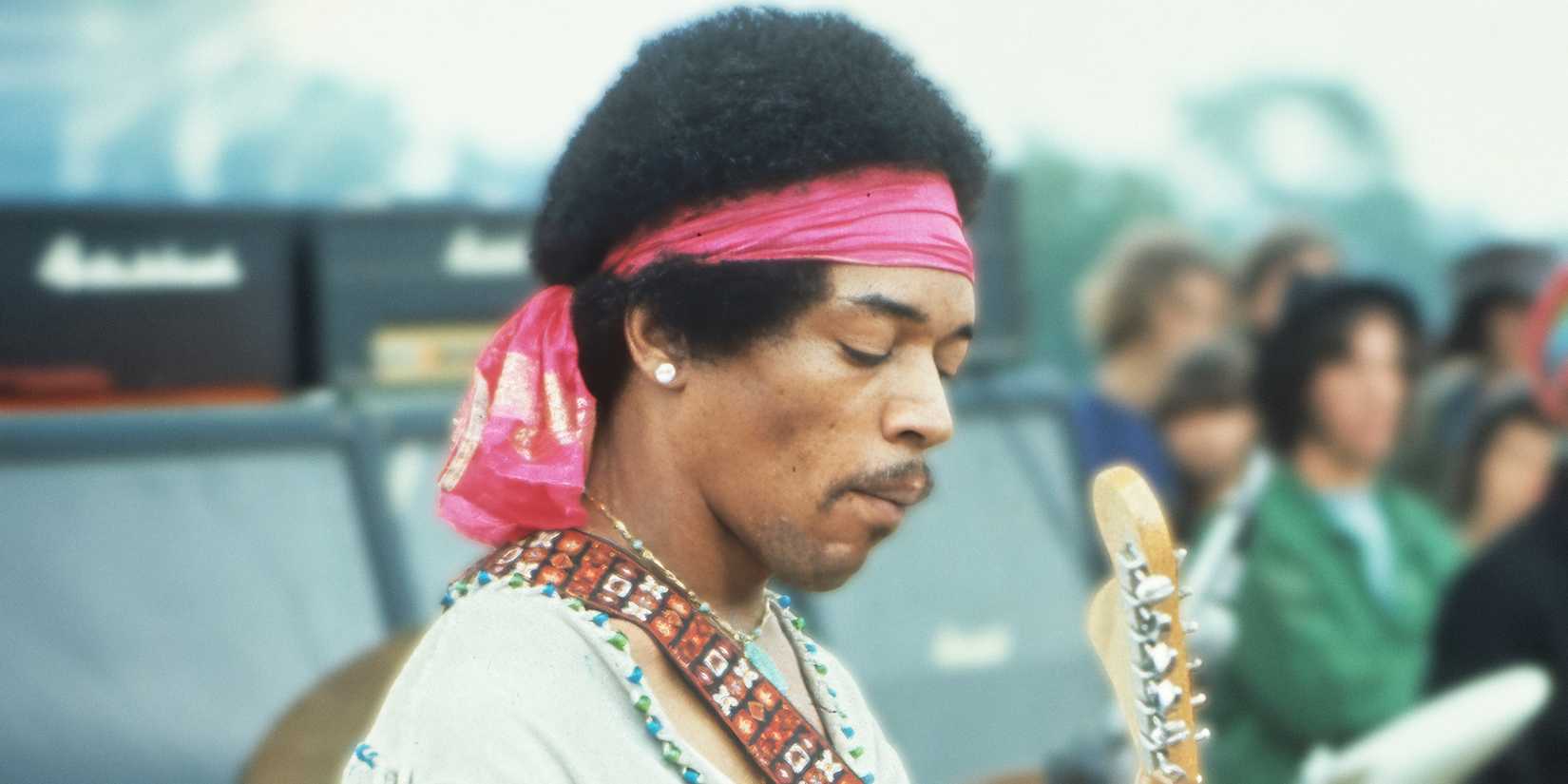 Jimi Hendrix looking down and playing his guitar while wearing a pink headband.