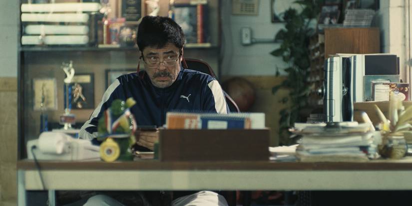 Benicio del Toro looks up from a cluttered desk in One Battle After Another