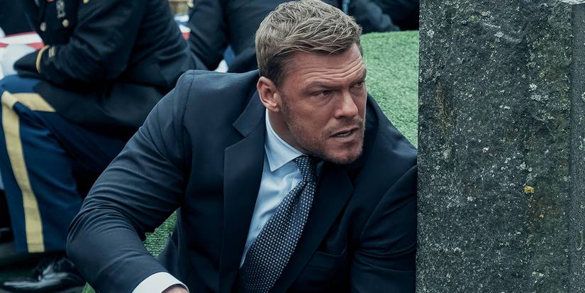 Alan Ritchson as Reacher hiding behind a tombstone as a funeral falls under attack