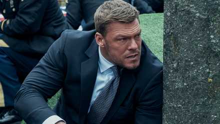 Alan Ritchson as Reacher hiding behind a tombstone as a funeral falls under attack