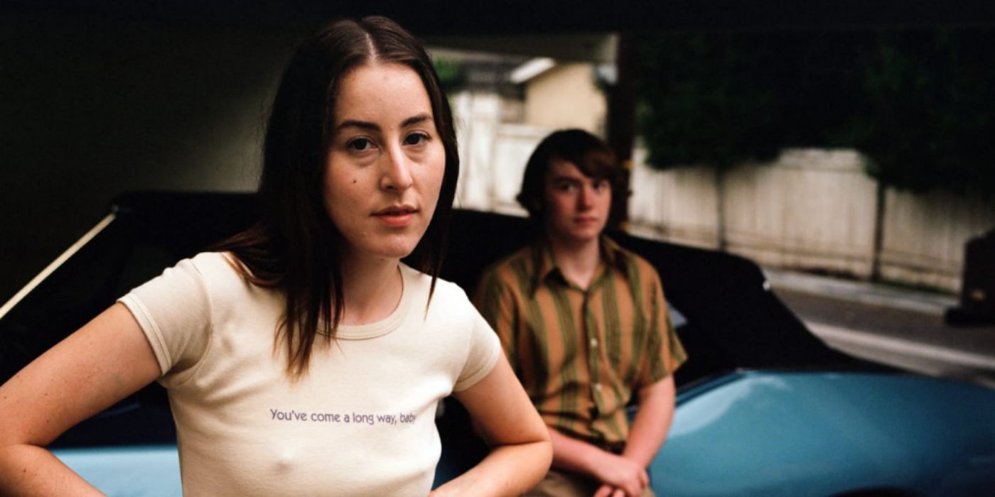 Alana Haim as Alana wearing a white shirt standing in front of a blue car in 'Licorice Pizza'