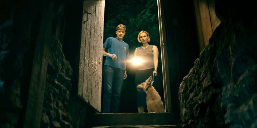 Mae Martin as Alex and Sarah Gadon as Laura with a dog shine a flashlight down into a cellar in Wayward.