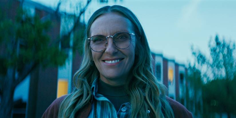 Toni Collette as Evelyn Wade wearing glasses and smiling outside a building at dusk in Wayward.