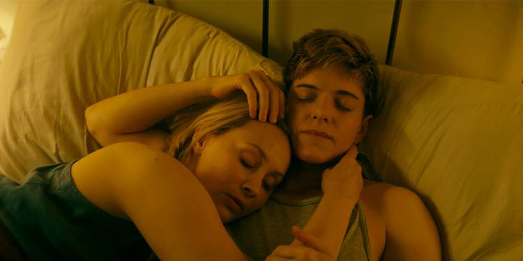 Mae Martin as Alex and Sarah Gadon as Laura cuddling in bed in Wayward.