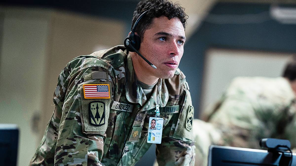 Major Daniel Gonzalez leaning on a table in a control room with a headset on, looking up in concern.
