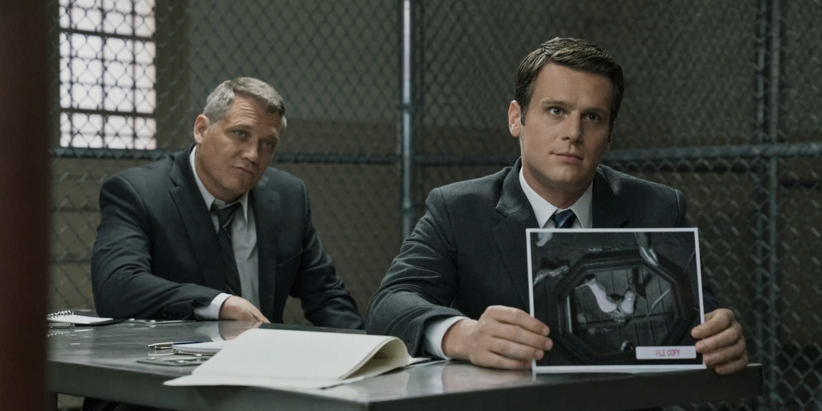 Holt McCallany and Jonathan Groff show a crime scene photo to someone off-screen in Mindhunter.