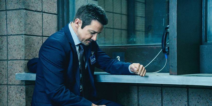 Mike sits at a prison telephone with his head hung, his arm propped on the counter with his fist clenched.