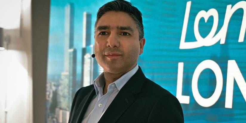 Zafar Jaffrey (Nick Mohammed) with a microphone in front of a blue sign in 'Slow Horses'