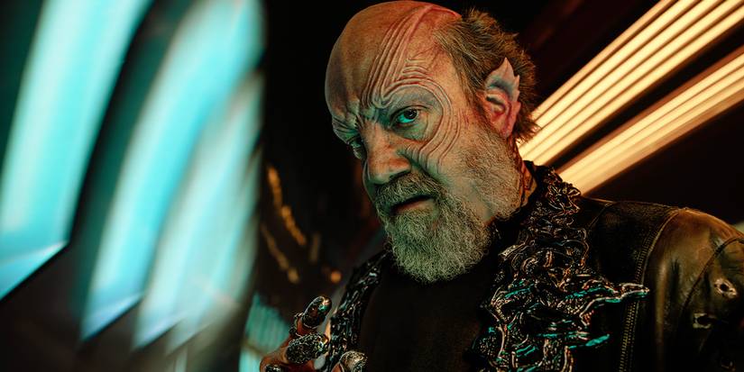 Close-up of Nus Braka in a gold embroidered outfit, pointing toward the camera in Season 1.