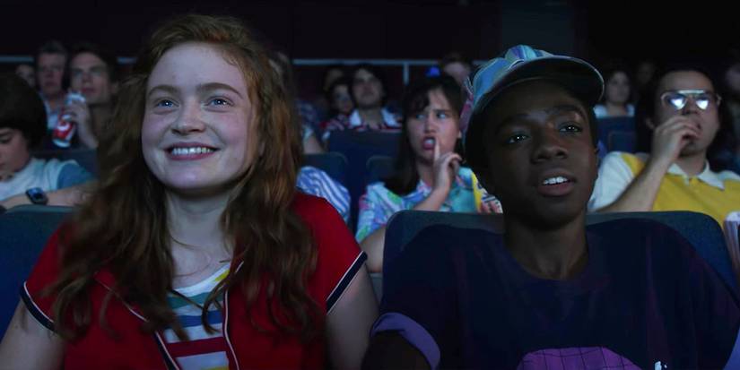 Max Mayfield and Lucas Sinclair in Stranger Things Season 3 sat in the Starcourt Mall Movie Theater facing forwards to the screen.