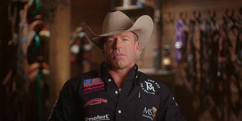 Taylor Sheridan in a shirt with brand logos and a cowboy hat looking at the camera in The Last Cowboy.