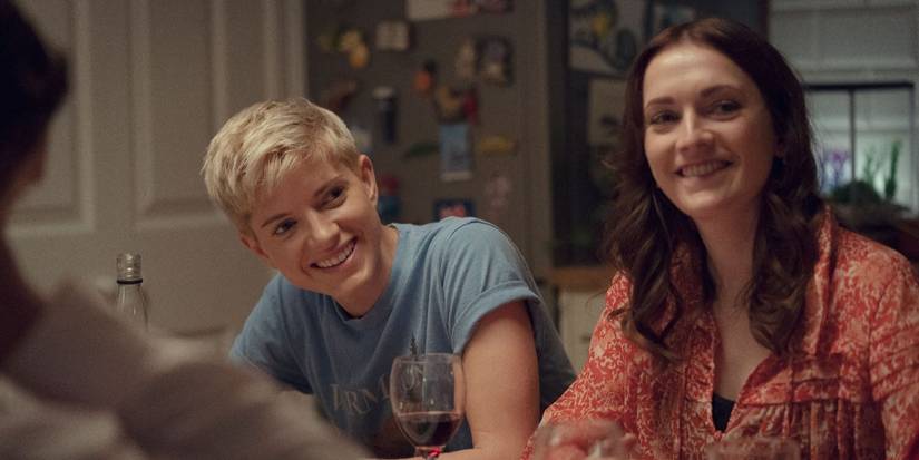 Mae Martin and Charlotte Ritchie smiling and looking to the side at a dinner table in Feel Good.