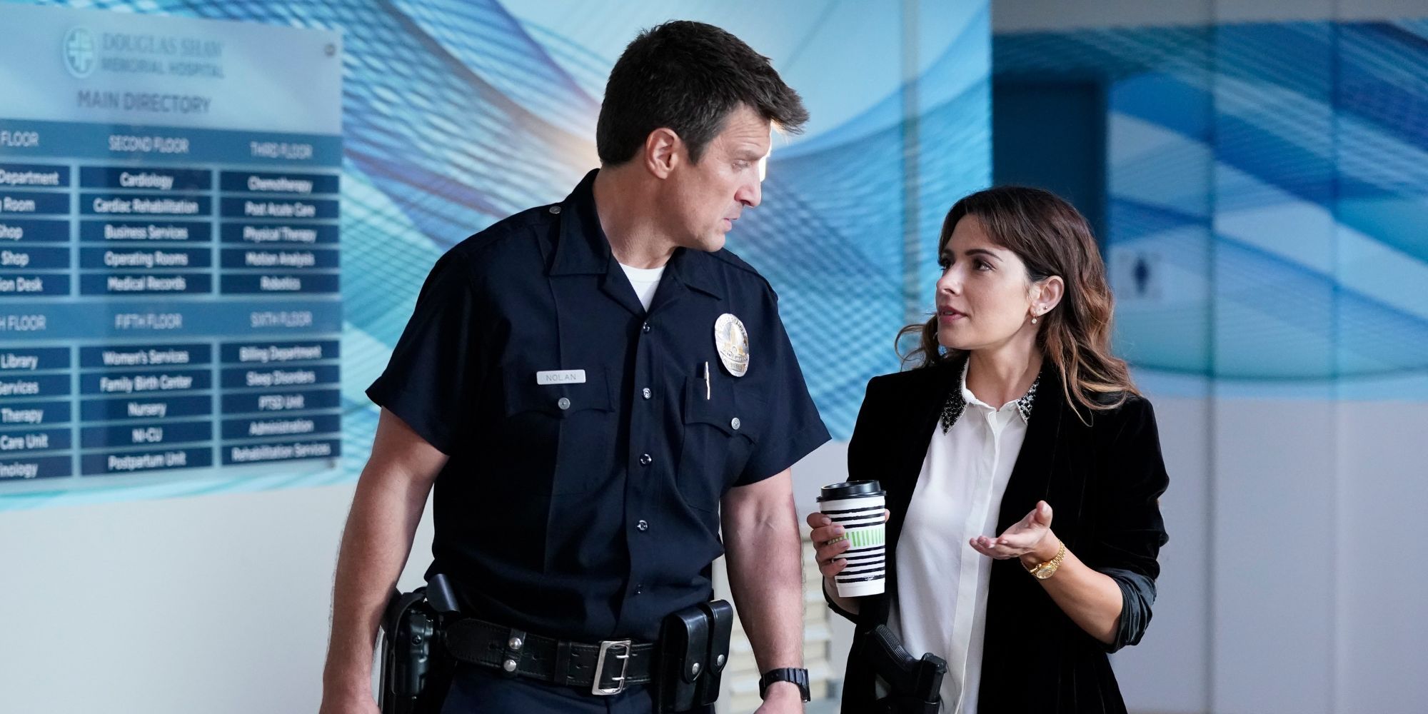 Jessica Russo (Sarah Shahi) chats with John Nolan (Nathan Fillion) as they walk down a hall in The Rookie.