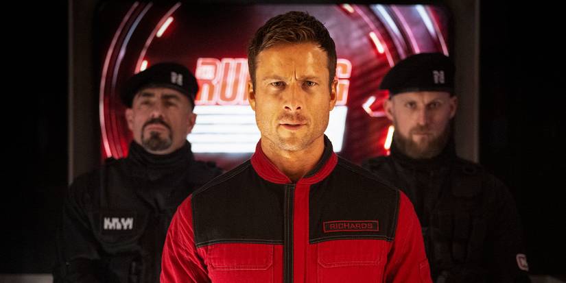 Ben Richards, in a red and black suit, frowning at the camera, flanked by two guards in The Running Man.