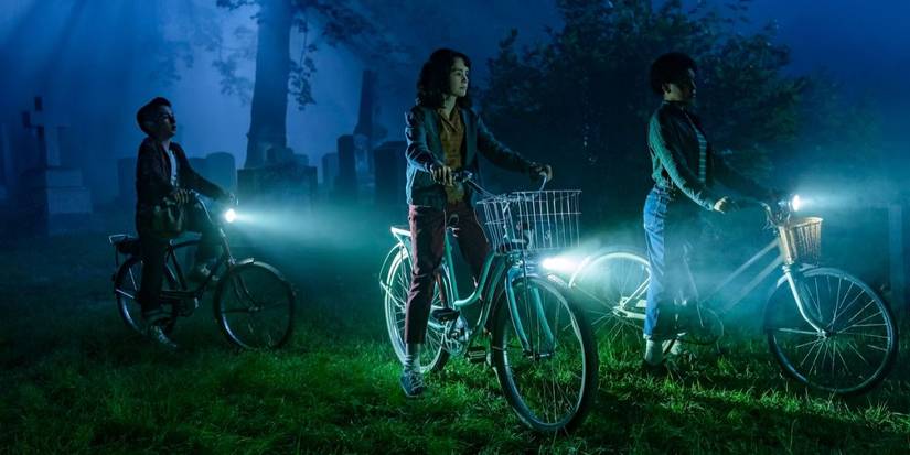 Rich (Arian S. Cartaya), Lilly (Clara Stack), and Ronnie (Amanda Christine) ride their bikes through a cemetery at night in 'IT: Welcome to Derry'