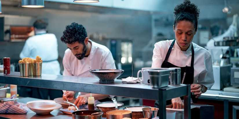A male and a female chef are preparing their dishes in a professional kitchen in the Boiling Point TV series.