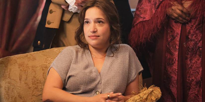 Abby sits on the couch smiling up at someone off-camera in Ghosts Season 5, Episode 5.