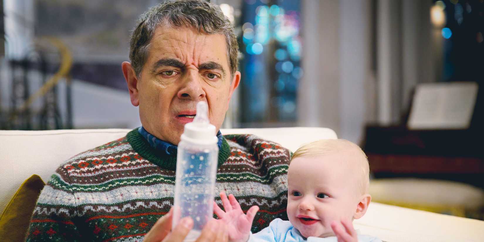Rowan Atkinson holding a baby and a baby bottle with a confused expression in Man Vs Baby
