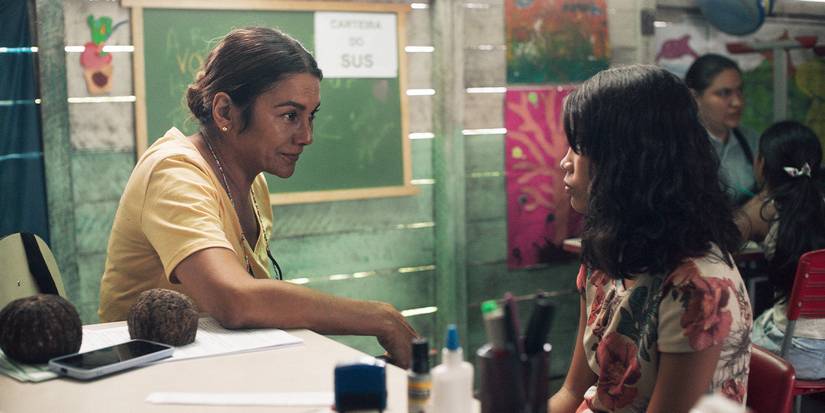 Dira Paes and Jamilli Correa sitting at a classroom desk talking in Manas.