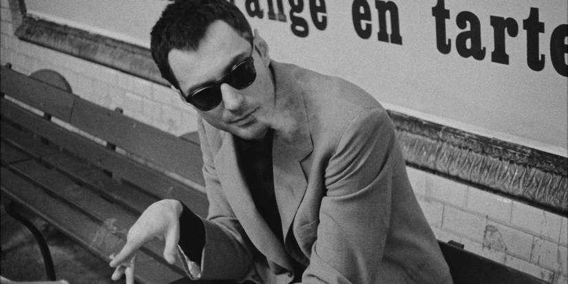 Jean-Luc Godard sitting on a bench and smoking a cigarette in sunglasses.