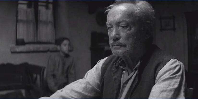 Udo Kier sitting on a table looking serious in Painted Bird