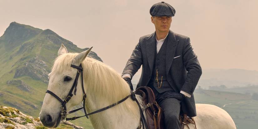 Tommy Shelby (Cillian Murphy) sits atop a white horse in the English contryside wearing a grey suit and his flat cap in 'Peaky Blinders' (2013-2022).