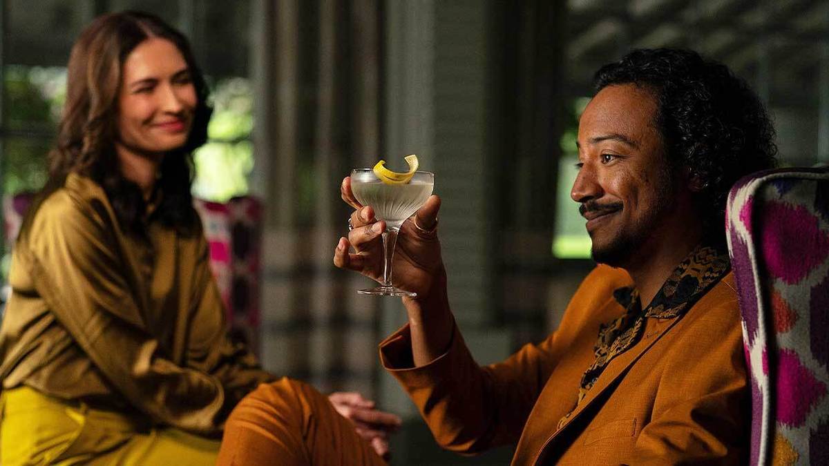 Karolina Wydra is smiling in the background in Pluribus while Samba Schutte smiles at a glass of cocktail in his hand.