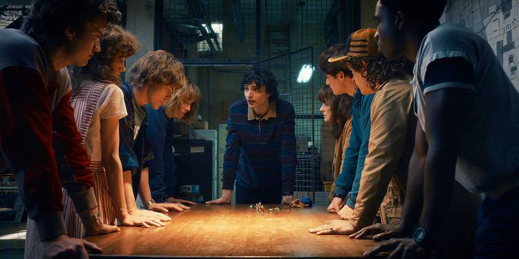 The cast of Stranger Things leaning in together around a table in Season 5.