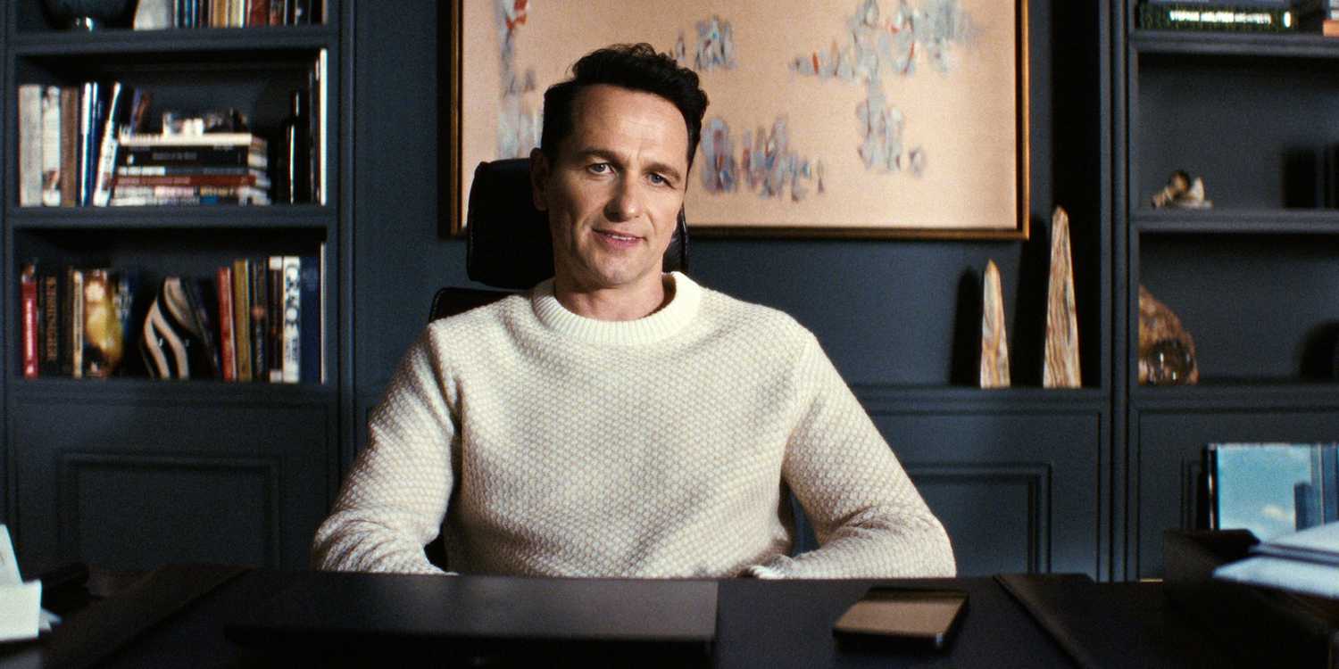 Matthew Rhys as Nile Jarvis sitting in a chair at his office desk with a smirk in The Beast in Me