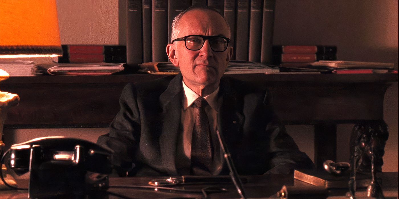 Don Licio Lucchesi (Enzo Robutti) sits behind his desk in a dimly lit room, wearing a suit and glasses in 'The Godfather Part III' (1990).