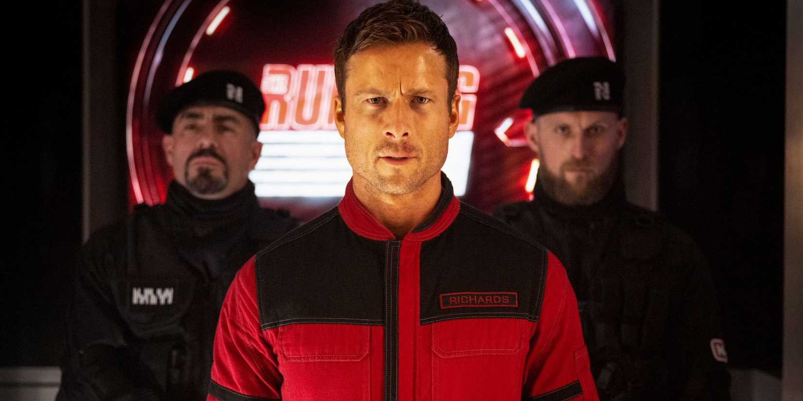 Glen Powell in a red suit stares ahead intently with two men in uniforms behind in The Running Man.