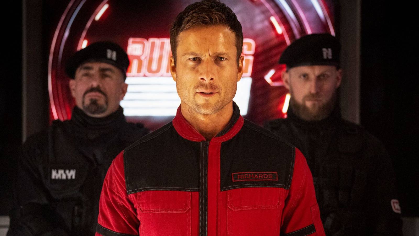 Glen Powell in a red suit stares ahead intently with two men in uniforms behind in The Running Man.
