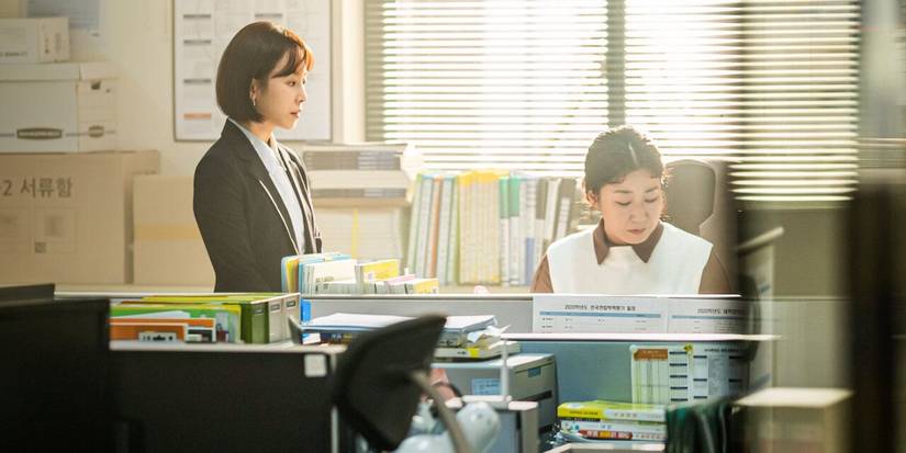 Seo Hyeon-jin standing besides Ra Mi-ran sitting at her desk in K-drama Black Dog