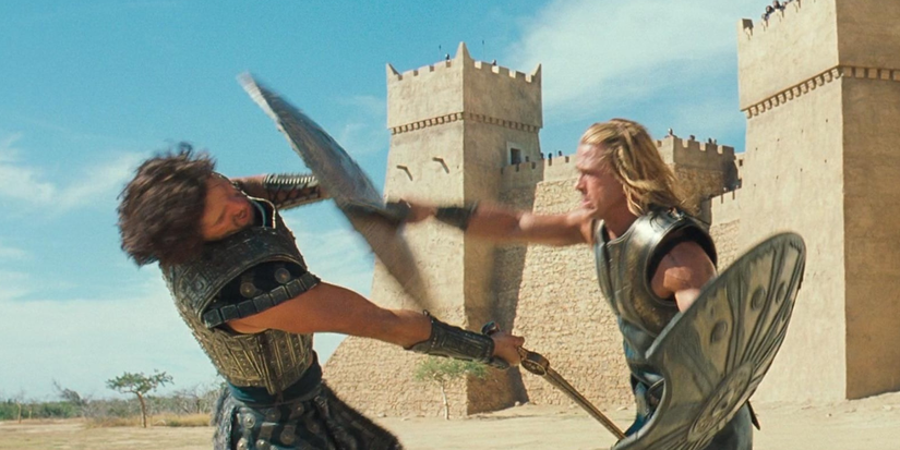 Two men carrying shields and swords fight each other in front of a fort