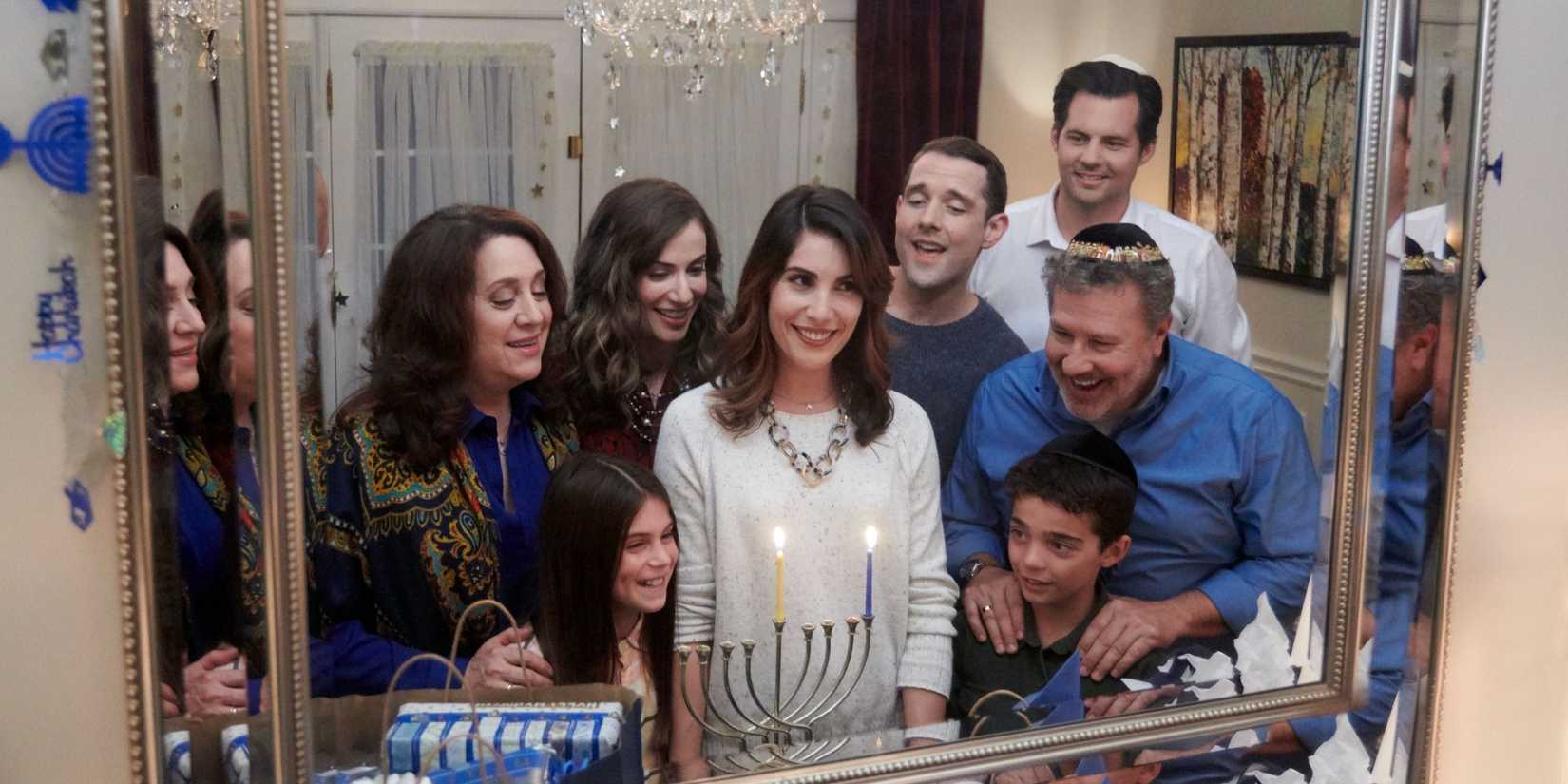 Carly Pope as Rebecca is reflected in a mirror with a menorah and several others smiling in Double Holiday. 